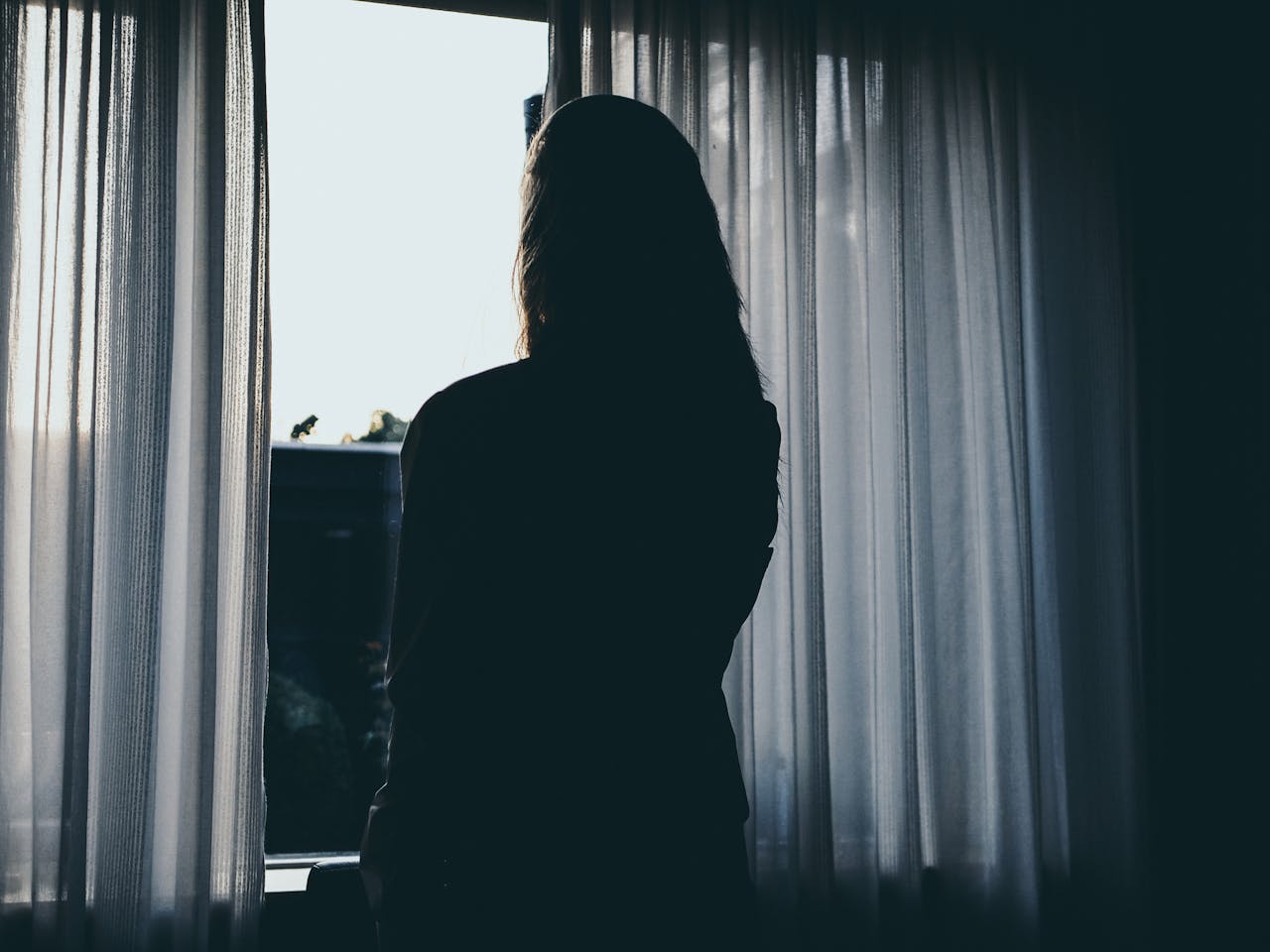 Silhouette of a woman standing by a window with flowing curtains, backlit by daylight.